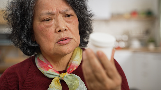 selective focus of confused asian senior woman with bad memory looking at a white medicine container in hand and can’t remember what it is. memory loss in Alzheimer’s concept