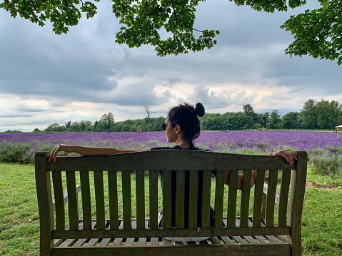 Young candid millennial girl is sitting on a bench in front of lavender fields with blossoming lavender flowers or walking between blooming lavender bushes.