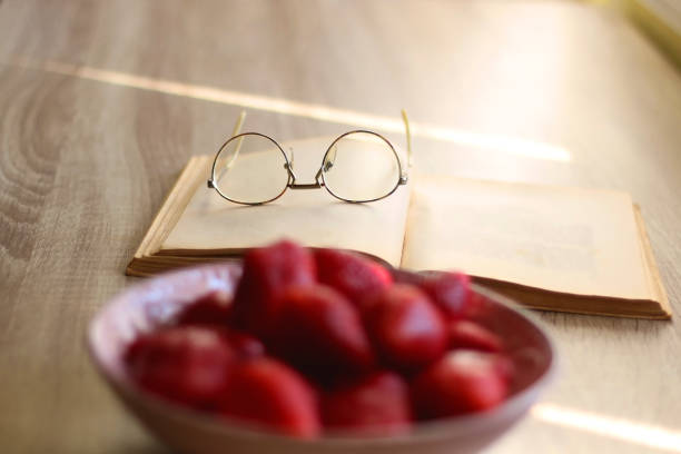 Bowl of fresh strawberries, open book and reading glasses on the table. Selective focus.