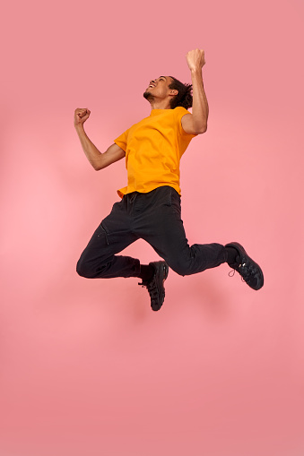 Full length shot of a cheerful funny black man in sneakers, crazy flying carefree jumping with raised arms, celebrating victory, having fun, rest, relax and leisure, isolated on pink background.