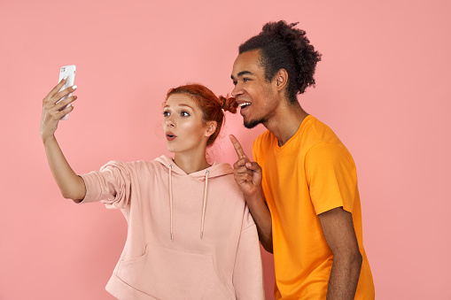 Studio shot of overjoyed dark skinned guy laughs sincerely while poses for selfie with ginger girlfriend, have fun together, use modern cell phone for entertaining, isolated on pink background.