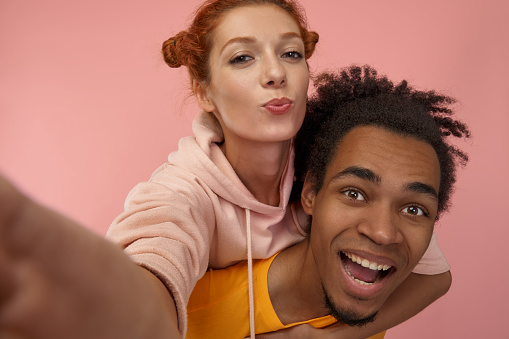Happy smiling mixed-race couple having fun and taking selfie on smartphone enjoy, piggyback ride, lovely woman hugs boyfriend from back, isolated on pink wall. People, fun, leisure time, valentine day