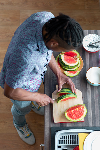 A directly above shot of a young man standing in the kitchen, he is slicing some fresh watermelon.