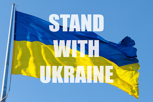 The national flag of Ukraine flutters in the wind and text STAND WITH UKRAINE. The concept of protecting Ukraine from Russian aggression. War in Ukraine