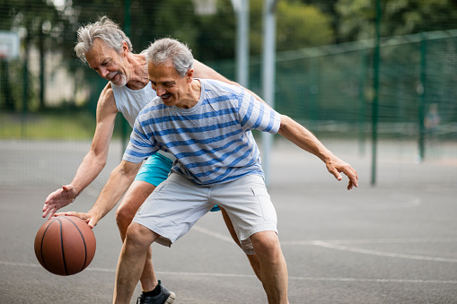 Two senior men playing basketball together at a basketball court in Newcastle-Upon-Tyne, England. One man is bouncing the ball on the ground while another man tries to steal the ball off him.