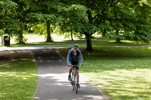 A senior man riding his bike on a footpath through a public park in Newcastle upon Tyne, England. He is looking at the camera and riding towards the camera.