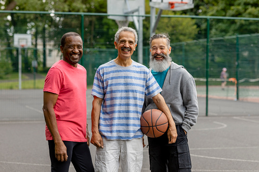 A portrait of three senior men standing outdoors on a basketball court in Newcastle-Upon-Tyne, England, they are looking at the camera and smiling. One man is holding a basketball on his hip.
