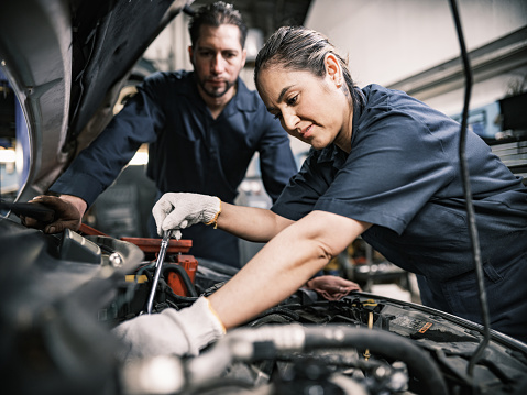 Mexican husband and wife mechanic team  in their shop. Mid adult man with beard and short hair and mid adult woman with braided hair wearing work clothes. Interior of car and motorcycle repair shop during day.