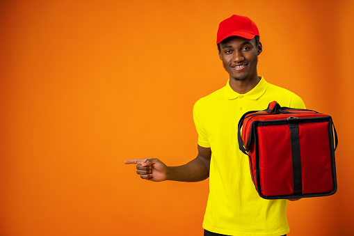 Smiling black male courier wearing uniform holding box in orange studio, close up