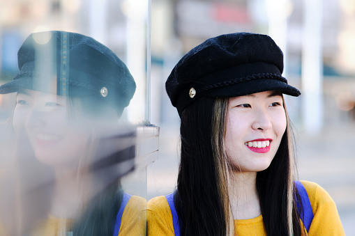 Portrait of a young woman wearing a hat or beret smiling looking away near glass at day.