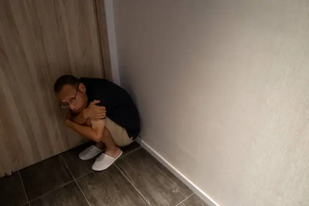 Asian man squatting on the floor in the corner of the room, very scared holding his knees