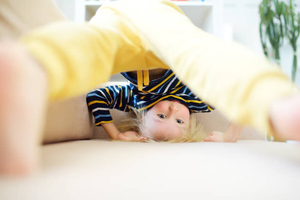 Little boy is standing on own head on sofa at home. Bored lonely child. Difficulties of family with preschool children during working at home. Baby having fun and standing upside down. stock photo
