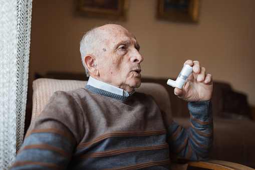 Elderly man sitting at home and using asthma inhaler due to his allergy issues