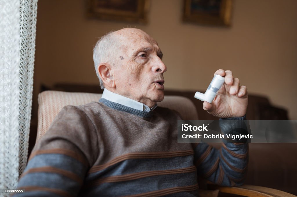 Old man having respiratory system issues and using an inhaler Elderly man sitting at home and using asthma inhaler due to his allergy issues Respiratory Disease Stock Photo
