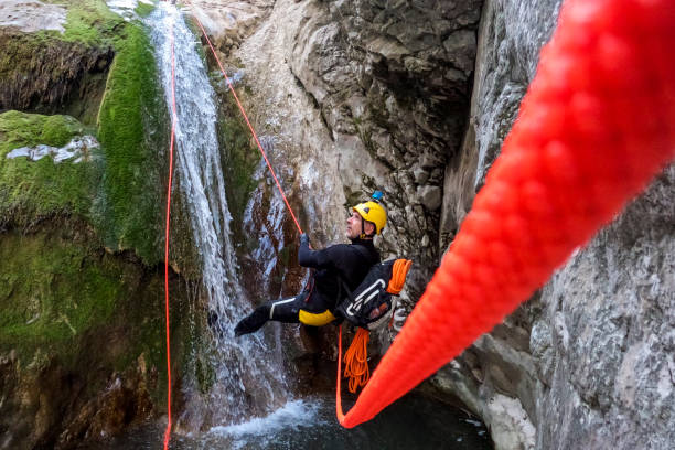 Male extreme canyoning team member seen in a spectacular canyon while rappelling down the waterfall with a neon orange rope during an adventure in the untouched nature.