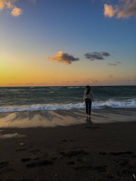 silhouette of a man standing on the beach watching the sunset.  you can see the reflection of the sun setting orange in the sky and sea surface