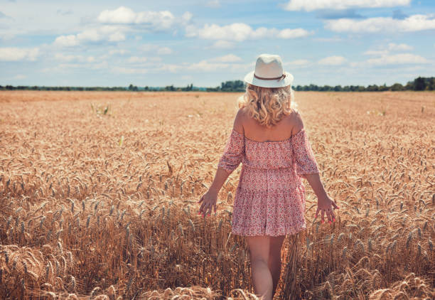A girl in a dress and hat lies on the ground in a wheat field and smiles, looking at the camera.