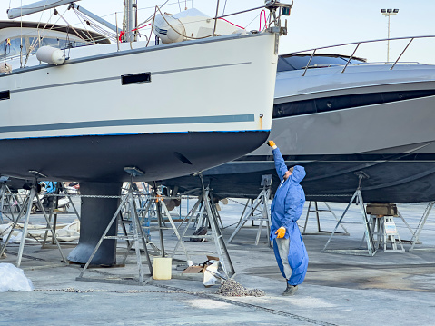 Shipyard worker with blue protection wear painting sail boat hull. Professional occupation and service at shipyards. A private sailing yacht has been lifted for maintenance and painting works at Turgutreis Marina in Bodrum. Close-up on employee.