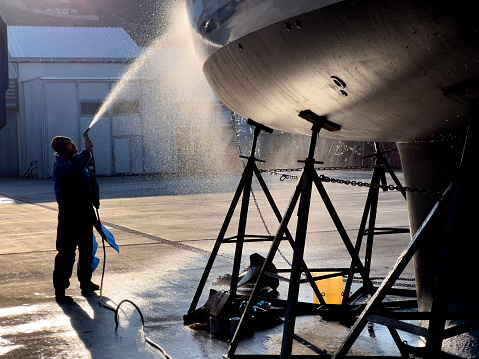 Blue collar worker spraying water on the bottom of private boat. Beautiful sun light reflecting on water drops. Yacht lifted for maintenance and painting works on dry dock. Professional occupation and service at shipyards.