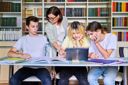 Group of teenage students studying in library class with female teacher. Mentor helping schoolchildren sitting at desks. High school, education, adolescent, back to school, back to college concept