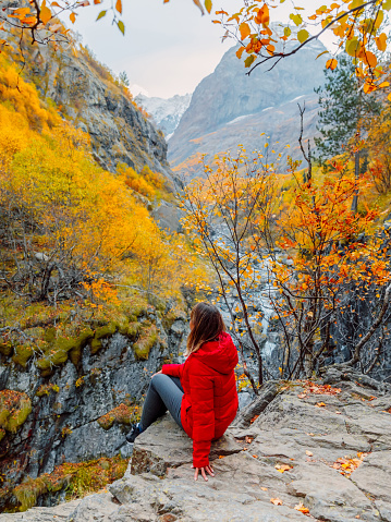 Rocky mountains and autumnal fall trees and woman in red jacket sit on rock. Mountain landscape with river