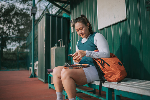 Asian Chinese mid adult woman sitting on bench in tennis court checking on her smart phone tracking her heartbeat rate after the game