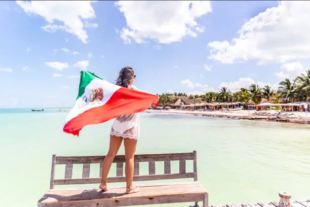 Woman stands on a wooden bench holding Mexican flag looking towards seaside with beach, palms and houses.