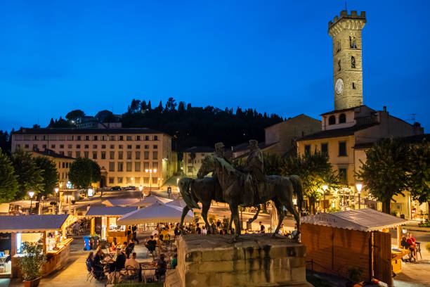 People sitting at the outdoor tables in Piazza Mino da Fiesole, the core of Fiesole. In foreground stands the monument 'Incontro di Teano', a bronze work by Oreste Calzolari (1906).