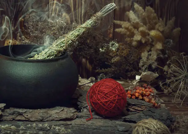 Magic items. Smoking twist of bitter wormwood herb for fumigation in iron cauldron, a ball of red wool, pine bark and dry flowers on wooden background, dark key, selective focus. Paganism, purification, voodooo, shamanism, rustic witchcraft concept