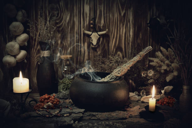 Authentic forest witch altar. Burning candles, dry herbs, iron cauldron, smoking mugwort, bulls skull mojo, bunch of garlic in wooden background in the dark, low key, selective focus. Authentic forest witch altar. Burning candles, dry herbs, iron cauldron, smoking mugwort, bulls skull mojo, bunch of garlic in wooden background in the dark, low key, selective focus. Pagan, Samhain, magic rite, voodooo, shamanism, occultism concept cauldron stock pictures, royalty-free photos & images