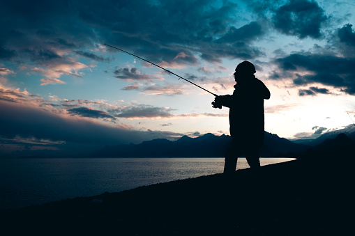 Silhouette of 30 year old fishing man fishing on the beach .The center of attraction is the world-famous Konyaaltı beach. The view of the Mediterranean Sea and Taurus Mountains, mountain ranges in winter. Closed air, sunset.The sun sets behind the mountains, the sea without waves.