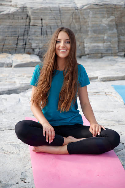 une jeune femme souriant à la caméra et assise en demi-lotus pose sur un tapis sur une plage de pierre - 7621 photos et images de collection