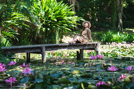 Portrait image of a beautiful young asian woman sitting on wooden bridge in a pond with pink lotus flowers