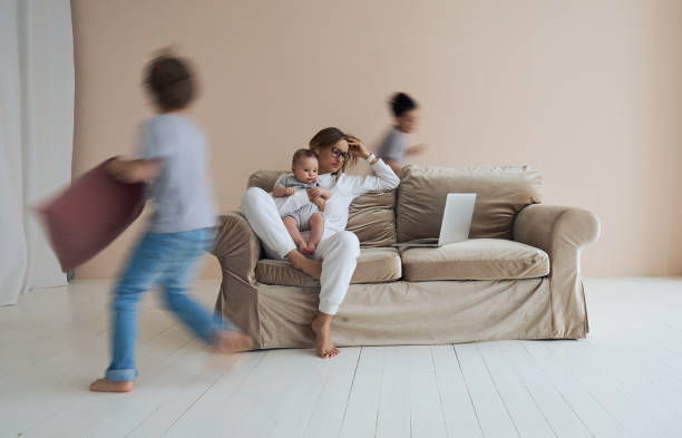 Young single mother working on laptop in loft sitting on couch while her sons running around her and shouting. stock photo