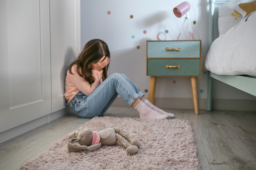 Unrecognizable sad girl sitting on the floor of her bedroom with plush doll lying. Selective focus on plush doll in foreground