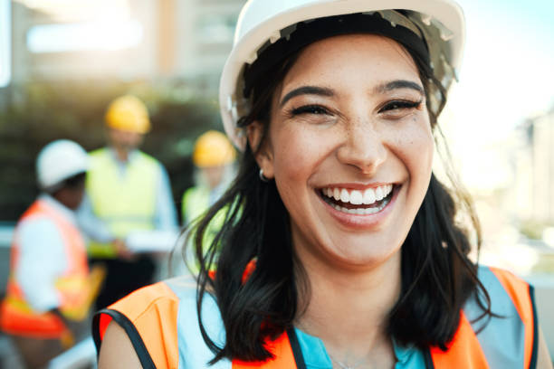 그녀의 동료가 그녀의 뒤에 일하는 동안 매력적인 젊은 계약자가 밖에 서 있는 샷 - construction safety protective workwear hardhat 뉴스 사진 이미지