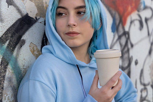 Teenage girl in light blue oversize hoodie with coffee to go and looking aside. Blue haired teen girl staying outdoors near graffiti wall. Cup mockup.  Hipster and adolescence concept