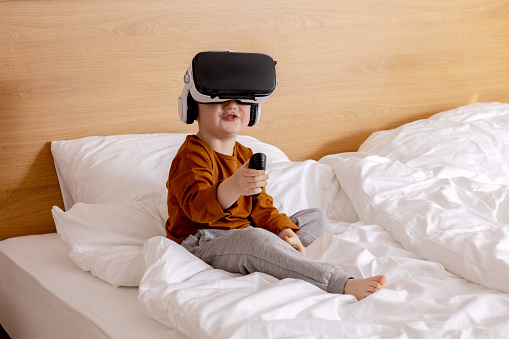 Pequeño niño adorable sentado en la cama en casa con auriculares VR y jugando videojuegos interactivos, explorando la realidad virtual. Lindo niño con gafas de realidad virtual. Futuro, gadgets, tecnología, educación en línea. photo