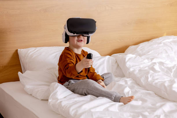 Little adorable boy sitting on bed at home with VR headset and playing interactive video game, exploring virtual reality. Cute child wearing VR glasses. Future, gadgets, technology, education online. Little adorable boy sitting on bed at home with VR headset and playing interactive video game, exploring virtual reality. Cute child wearing VR glasses. Future, gadgets, technology, education online digital native stock pictures, royalty-free photos & images