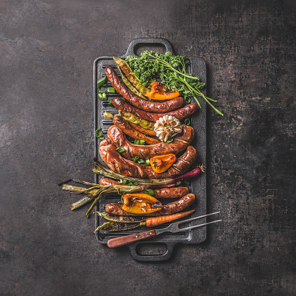 Grilled food on cast iron plate: sausage, pepper, spring onion, garlic, paprika, herbs, carrot with meet fork. Summer Barbecue concept on dark table. Top view