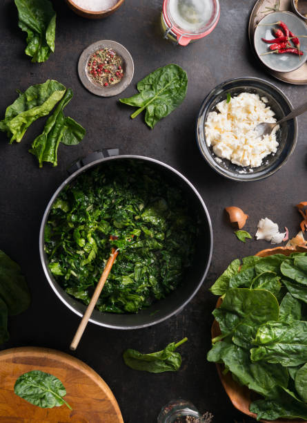 Spinach cooking preparation. Stewed spinach in black cooking pot with spoon on rustic background with ingredients and kitchen utensils stock photo
