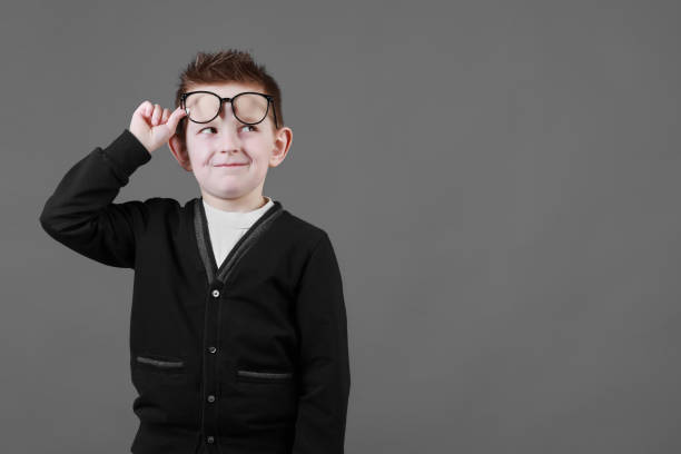 Smart child boy adjusts his glasses and having fun on the grey background, close-up. Studio juvenile portrait in casual clothes. copy space.