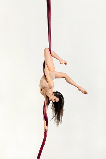 Slim female aerialist performing falling stunt on aerial ribbons while hanging upside down on white wall background