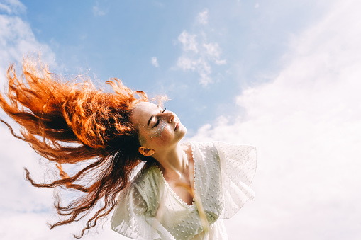 Young happy woman with flying long curly hair in sunshine. Sky background. Copy space.