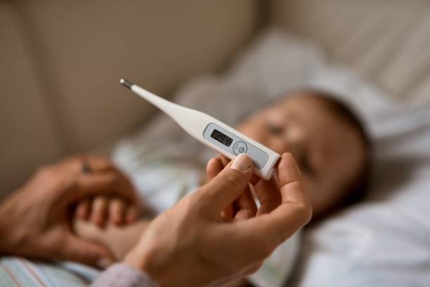 Worried young mother sitting on sofa beside her sick son with high fever. Mom measures temperature using thermometer of sick child lying under blanket at home stock photo