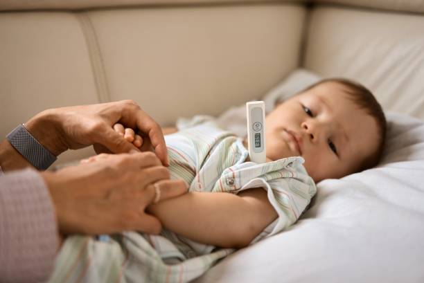 Worried young mother sitting on sofa beside her sick son with high fever. Mom measures temperature using thermometer of sick child lying under blanket at home stock photo