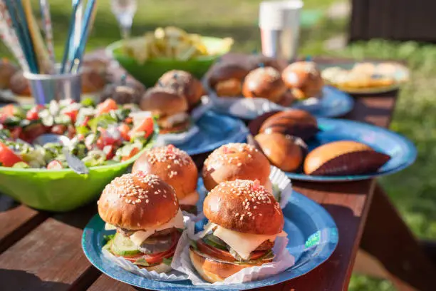 Photo of Buffet table with many mini burgers at outdoors birthday party for children, kids. Catering at wedding day. Bright tasty hamburgers with fish, chicken, meat, salad. Dinner in country house in village