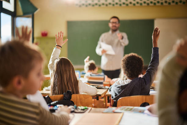 istock Back view of students raising their hands to answer the teacher's question on a class. 1388919896