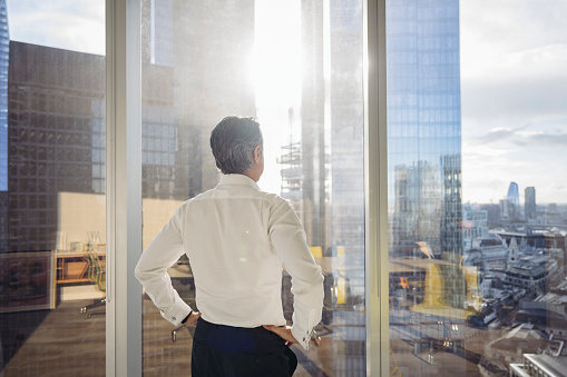 Grey-haired corporate executive in early 40s standing with hands on hips and looking out at London’s financial district in bright afternoon sunshine.
