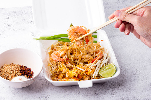 Hand holding chopsticks and pick up a prawn and noodle from Fried noodle Thai style with prawns in foam box, Pad Thai with vegetables. popular street food of Thailand.